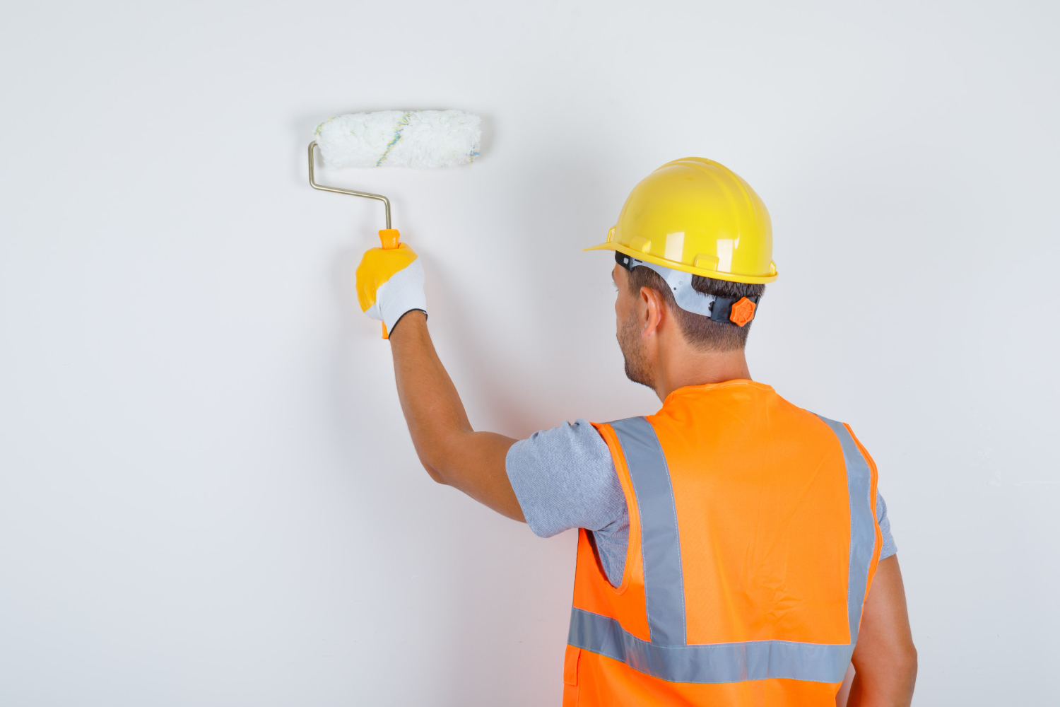 A male builder wearing an uniform, helmet, gloves: painting a wall with roller back